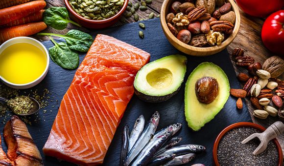 Image shows a variety of foods including a piece of salmon, avocados, sardines and two small bowls of nuts and seeds