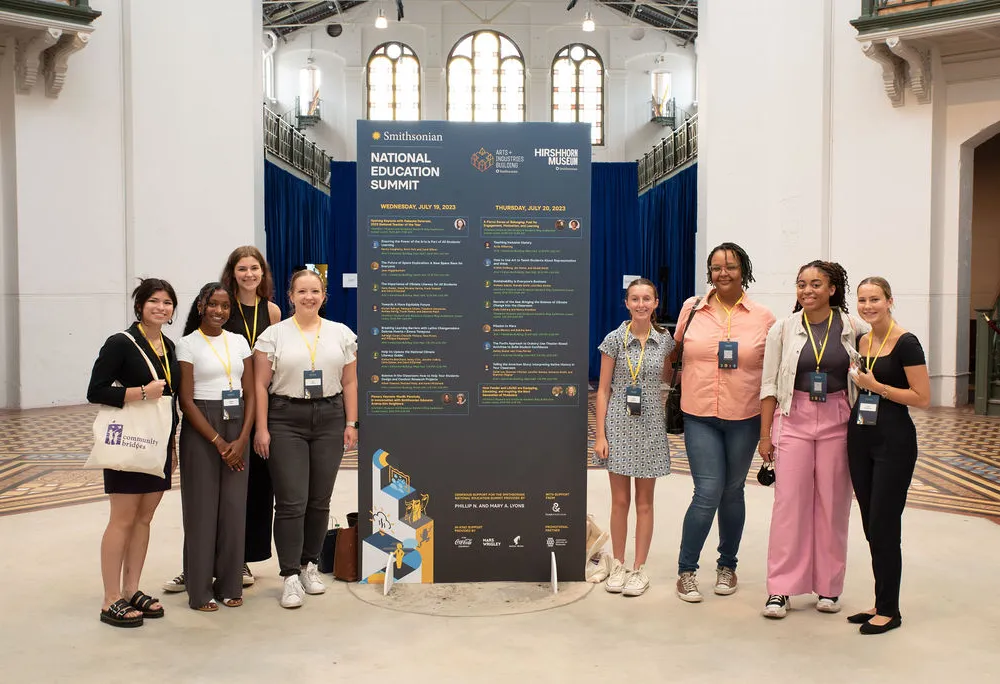 A group of teachers gather around a large blue billboard of the Summit schedule