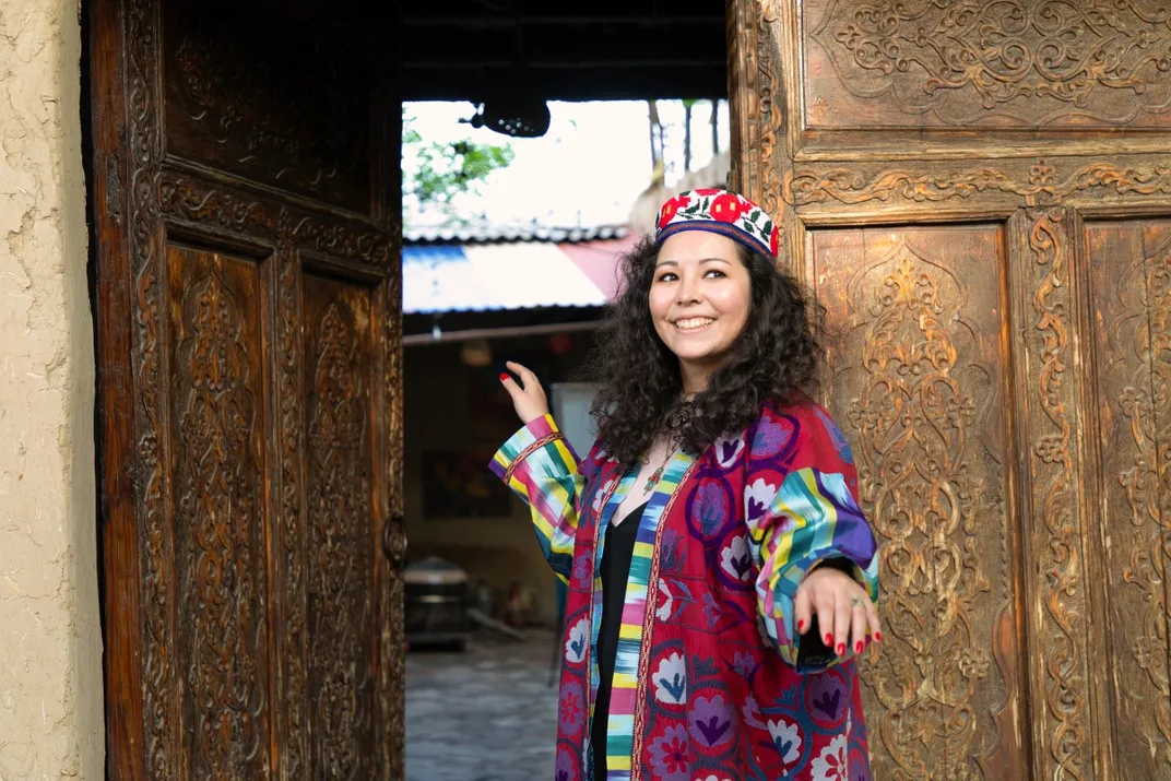 A woman stands in a doorway while wearing a long colorful garment and smiling to the right of the camera. She leans against a wooden door that has intricate designs.