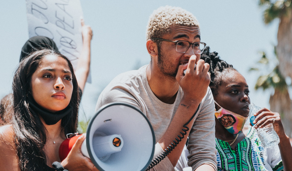 Three young activists are outside at a protest – looking off into the distance. They all appear to be African American. One is holding a bullhorn and has a determined look on her face. Another is holding the bullhorn’s microphone and speaking into it.