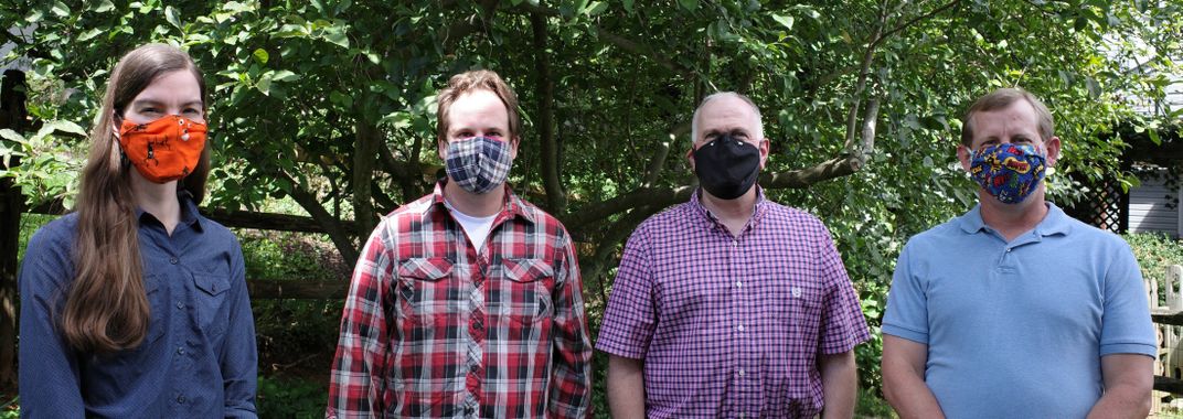 Four people, all wearing masks, stand in front of green leafy trees and smile a few feet apart from one another