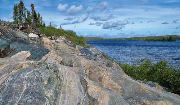 A slab of multicolored, sedimentary rock is pictured with vibrant trees and water surrounding it.