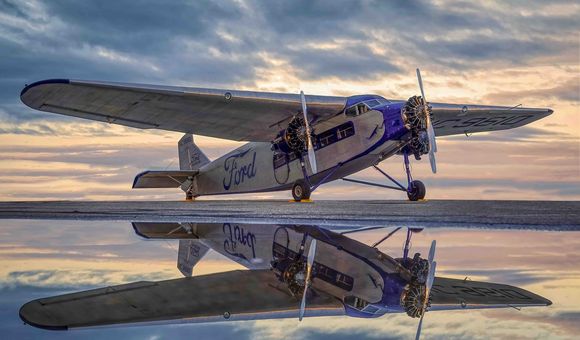 A vintage silver Ford Tri-Motor aircraft parked on a wet tarmac under a dramatic, cloudy sky at dusk. The plane features corrugated metal skin, three engines, and the "Ford" script logo in blue on the fuselage, all reflected in the water on the ground.