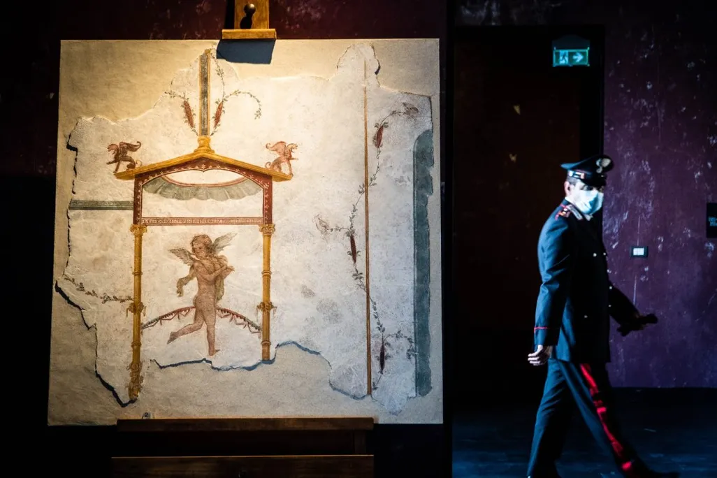 The recovered fresco fragment hangs on an easel display, lit up in foreground; in background, a police officer wearing a mask walks past the display