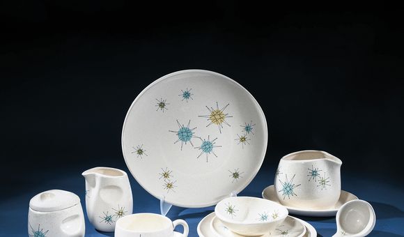 Photographed against a dark blue background is a studio image of a set of cream-colored ceramic dishware bearing a pattern of star-shapes in blue, yellow, and green. There are several pieces, including a platter, a gravy boat with a ladle, a creamer and s