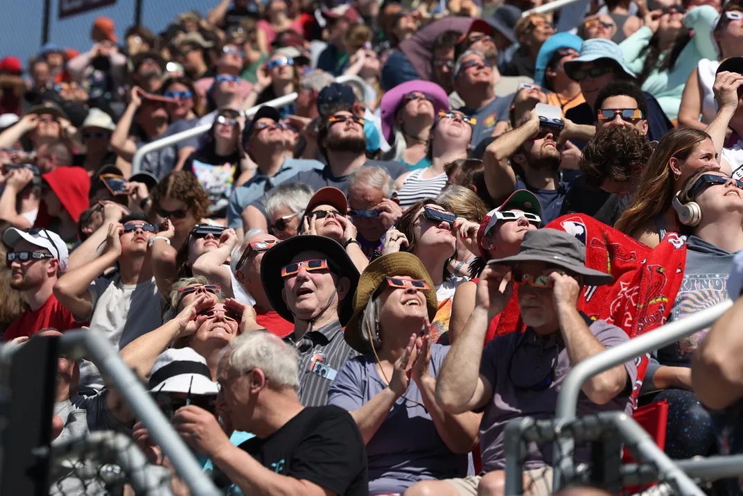 Crowds gather in a stadium wearing eclipse glasses