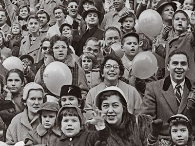 A crowd of people cheering and waving at a parade, circa 1955.