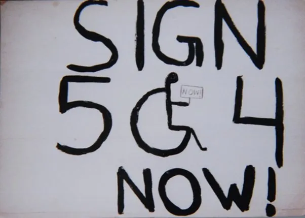 A handwritten sign with black letters on white background, which reads "Sign 504 Now!" The "O" is a person sitting in a wheelchair, holding a smaller sign that says "Now!"