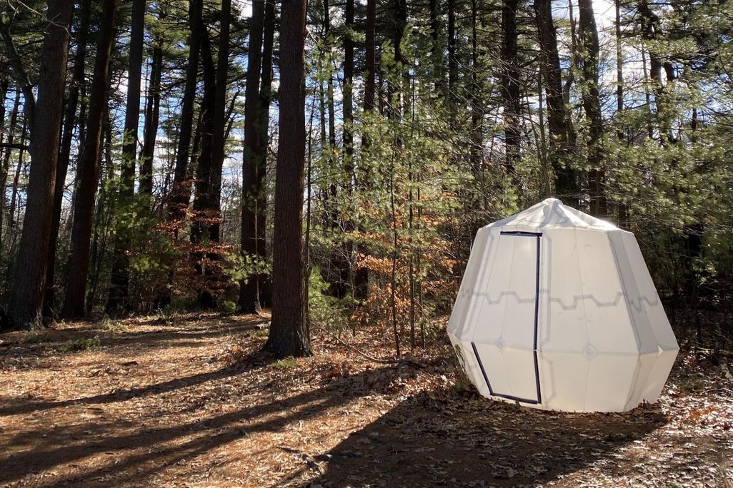 A white octogon-shaped origami tent sits in the woods in New England