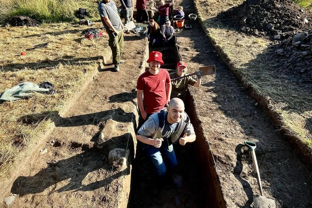 A group of people in a shallow ditch pose for a photo while digging
