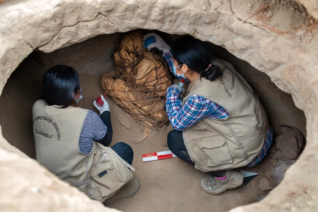 Two people kneel in an oval-sized opening and carefully brush the mummified body