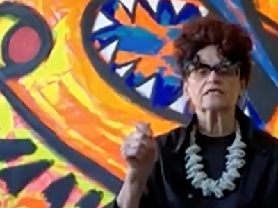 Detail of color screenshot of Judith Bernstein in front of one of her paintings wearing a black blouse and light-colored, chunky necklace holding her hand up in a fist as if making a point in conversation.