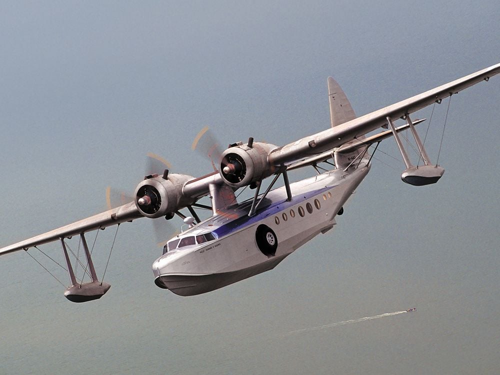 Silver twin engine flying boat over a vast ocean. A small boat leaves it's wake below.