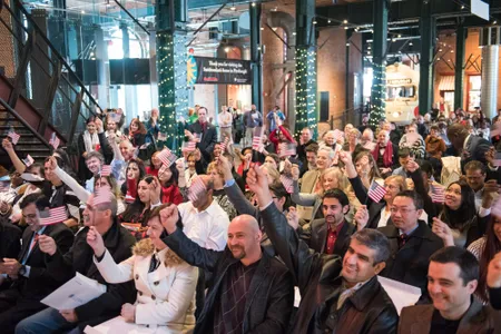 A group of people sit in chairs at the History Center raising mini American flags during a Citizenship ceremony