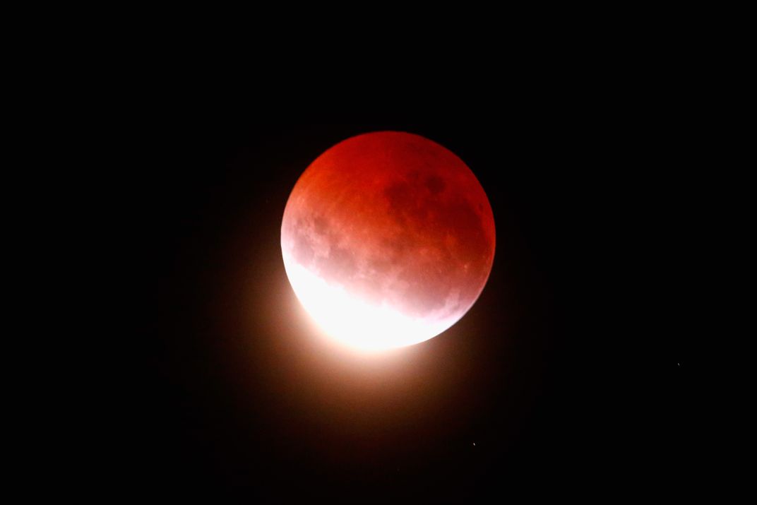A moon almost entirely eclipsed, with bright white light at its lower left curve and mostly reddish shadow covering the rest of its surface