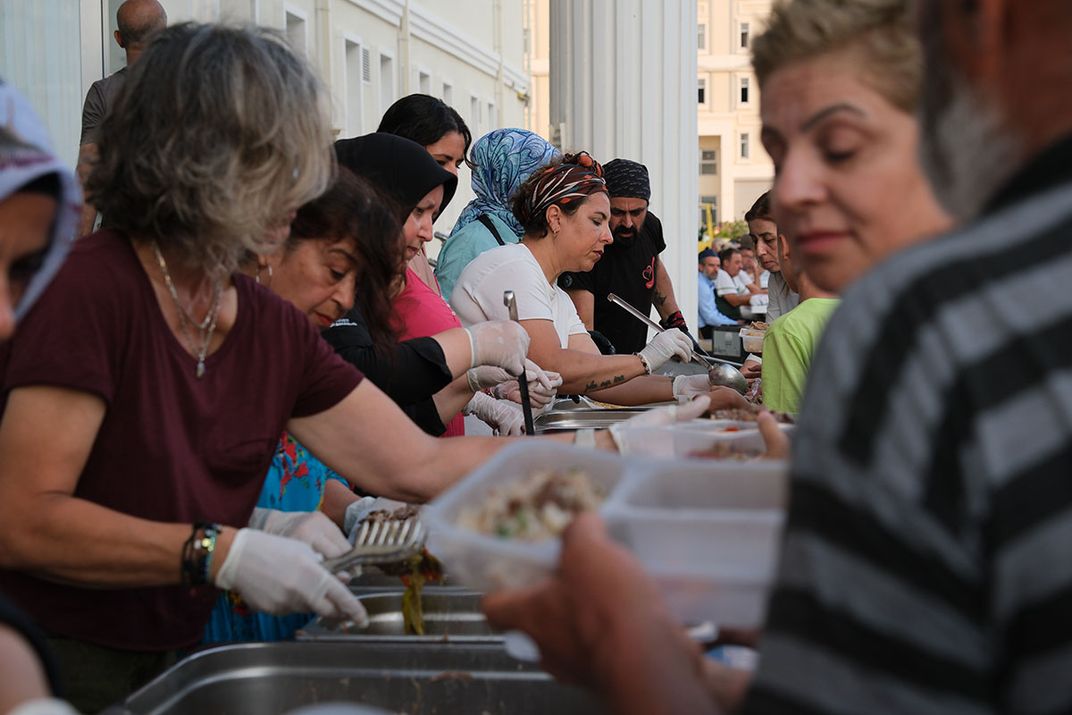 Outdoors, people line either side of a table serving and being served food into plastic trays.
