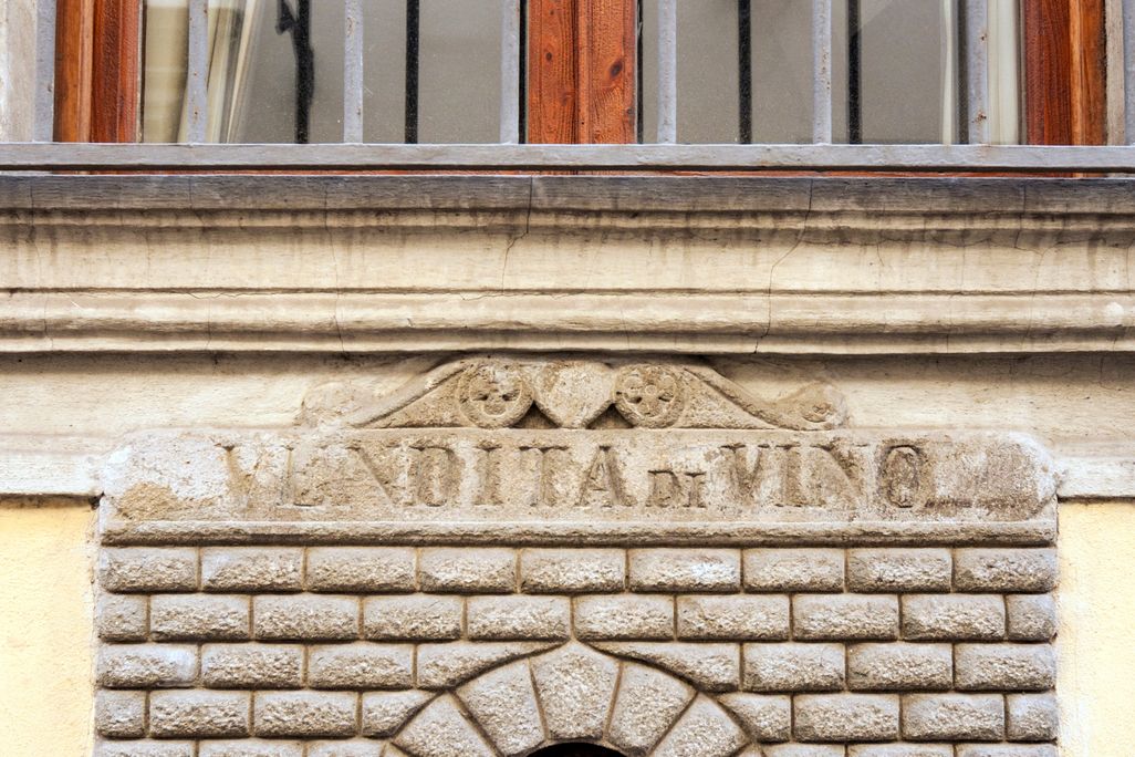 A small door cut into the side of a house. The door has engraved stone around its entrance, made to look like a tiny building facade. It bears an inscription that has worn away, but includes the word "VINO"