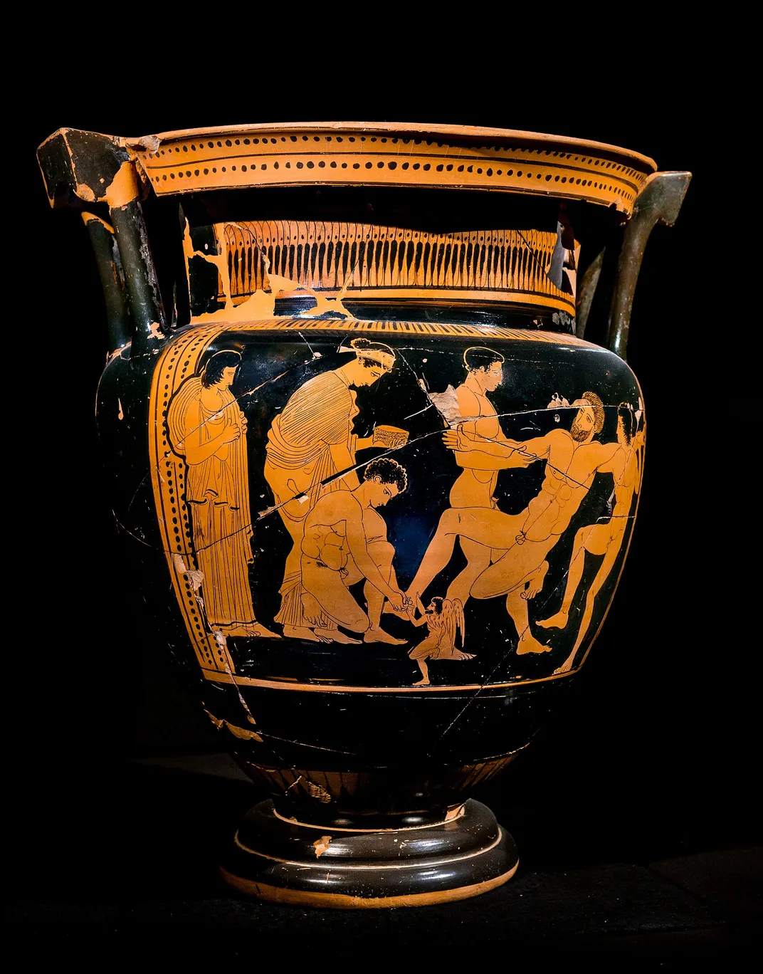Medea watches as Jason uses a tool to remove a bolt from Talos' ankle.