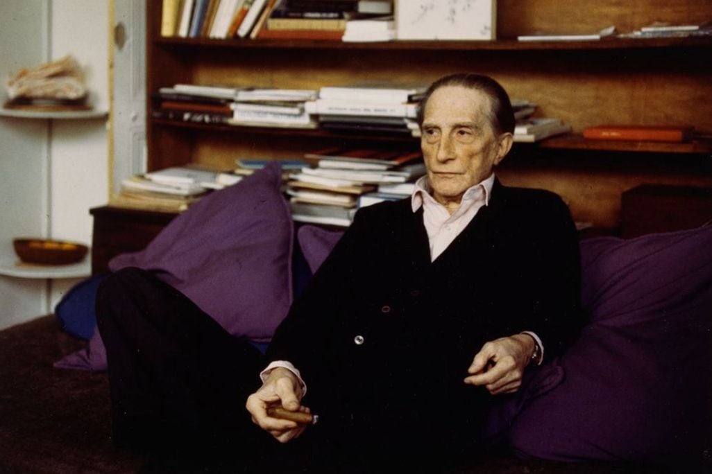 Elderly man in black sweater sitting in purple chair in front of wooden bookshelf