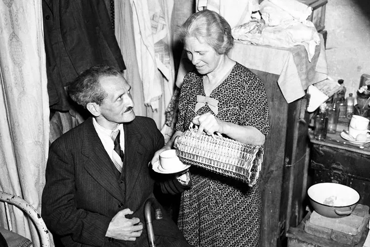 A white woman in a dress pours tea for a seated man in a suit, in a small home crowded with items (longform main)