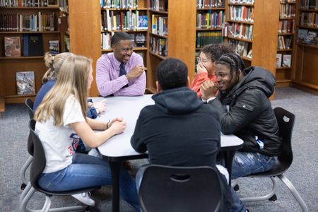Teacher Kurt Russell sits at a square table in the school's library with five students, smiling and in conversation.