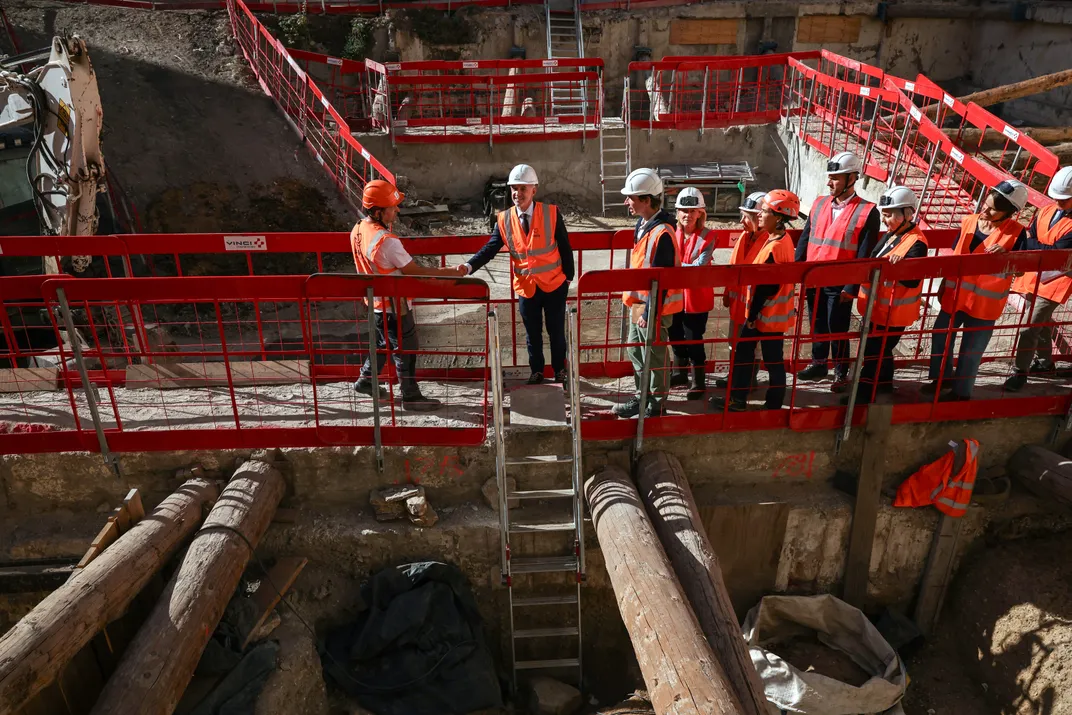People in hard hats and orange vests stand on a platform above an archaeological site