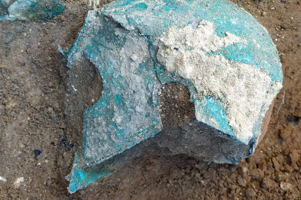 A view of a blue-green oxidized metal helmet, with sharp curved cheek plates that extend past the face, lying on the ground in the dirt