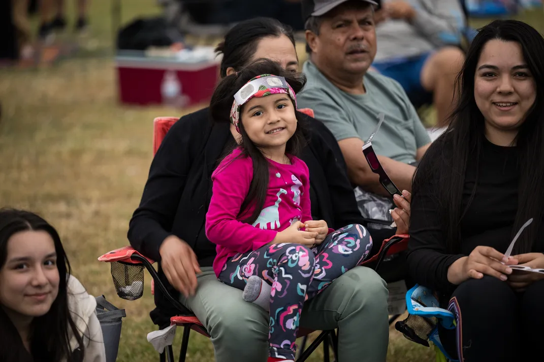 people including a young girl on a woman's lap sit smiling waiting for the total solar eclipse
