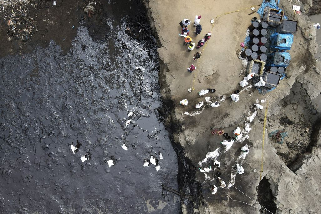 An aerial image of an oil spill off Peru's coast. The photo shows a contaminated beach smeared with black crude oil and workers in PPE trying to clean up the beach.