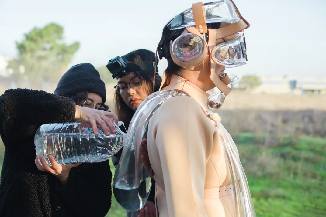 Two Latina women pouring water into a glass element on Tanya Aguiniga's suit, at the U.S./ Mexico border.