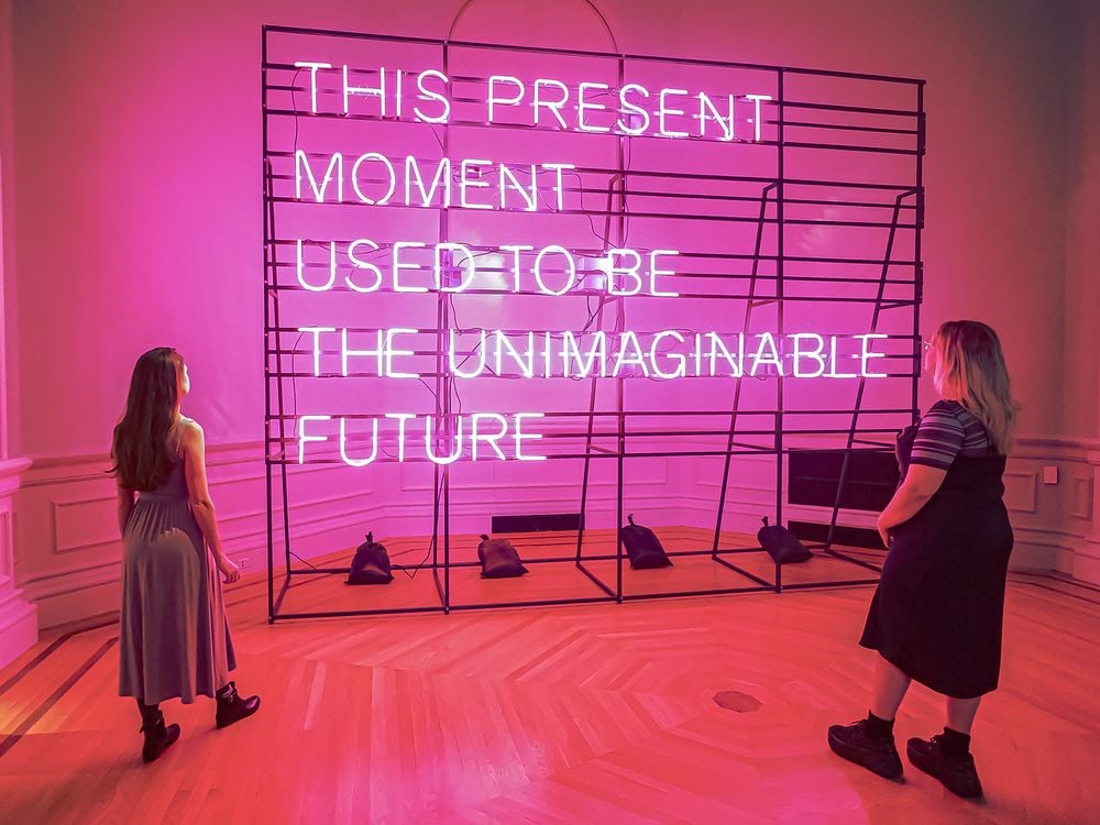 Two people stand in front of a pink neon sign that states "This Present Moment Used to Be the Unimaginable Future"
