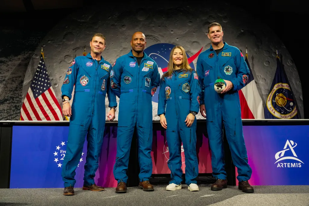 four astronauts stand in front of a press conference table wearing blue jumpsuits and smiling