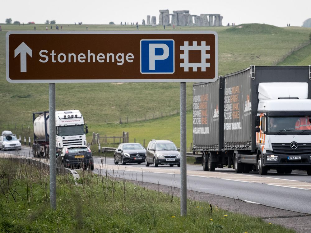 A busy road is in the foreground, with trucks and cars, and a large sign pointing to Stonehenge; the iconic stone structures are visible in the background