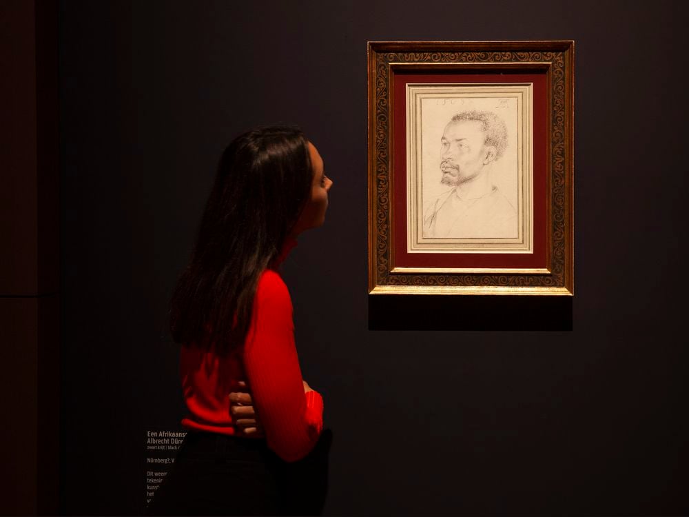 A person with long dark hair stands in a darkened gallery and looks at an illuminated white paper sheet, with a sketch of an African man's head and shoulders
