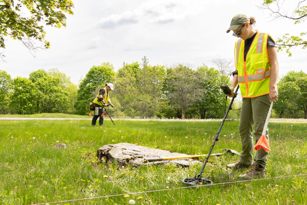 Two people wearing safety vests using metal detectors to scan the grass