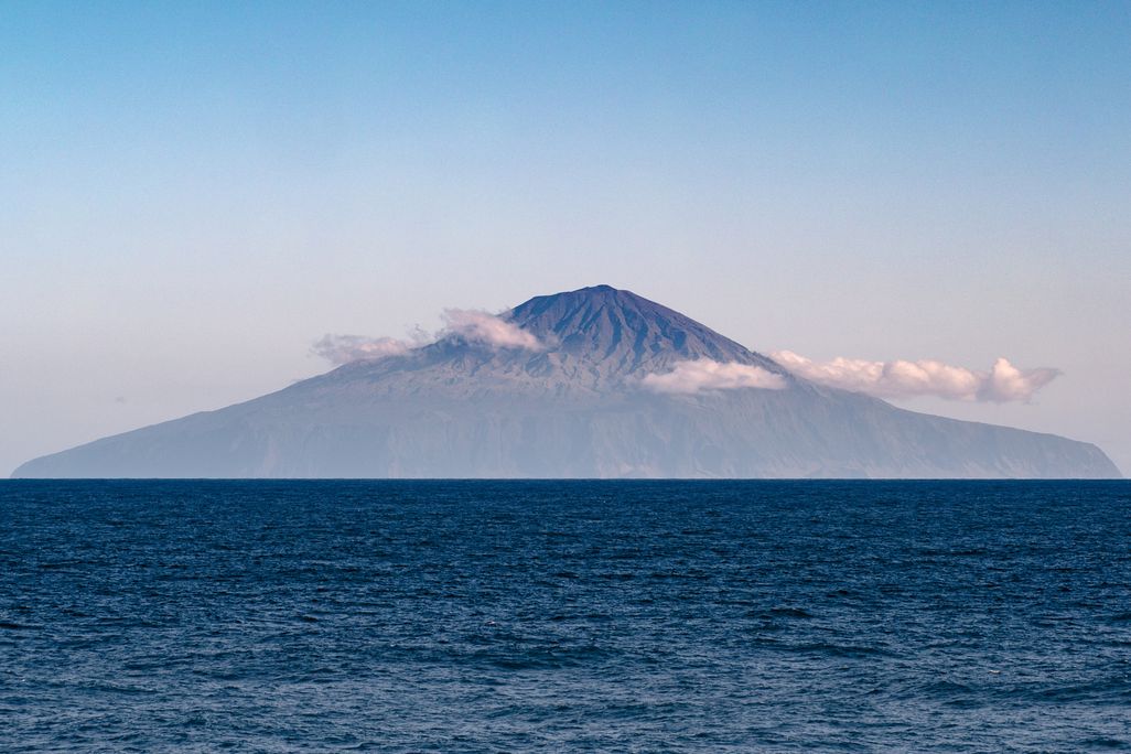A photograph of Tristan da Cunha taken from a boat out at sea. The blue ocean water meets the horizon halfway up the photo. The island, shaped like a flat triangle sits at the horizon. It has small clouds in front of it and a blue-pink sky in the backgrou