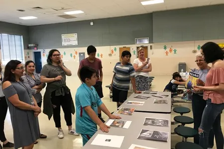 Group of adults and middle school children standing and actively discussing a set pf printed pictures laid out on a central table.