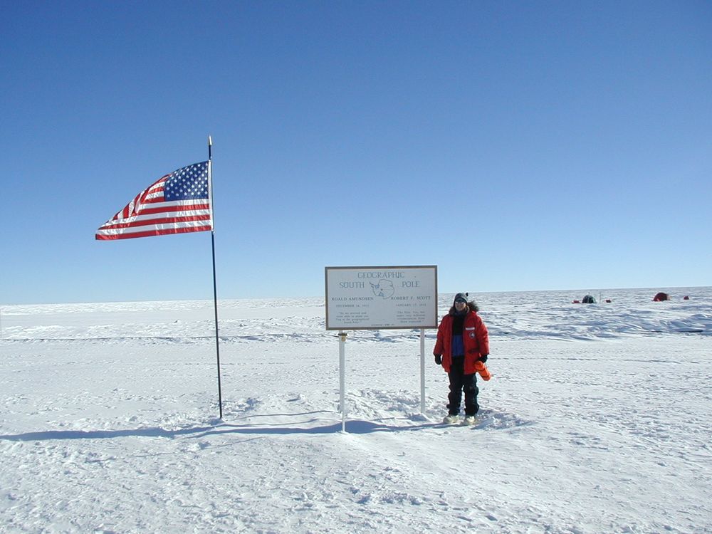 Geolgist Cari Corrigan poses for a picture in the snow next to an American flag and a sign that reads "Geographic South Pole."