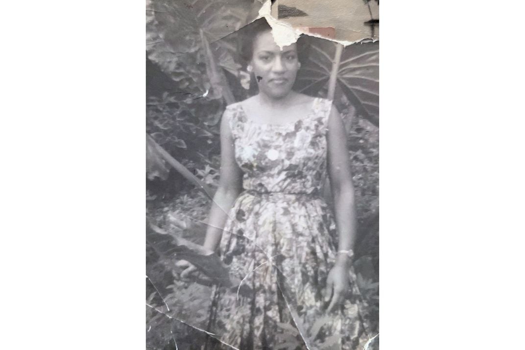 A woman posing among the wide leaves of a tropical plant, wearing a floral print dress. Black-and-white photo that is crumpled and torn.