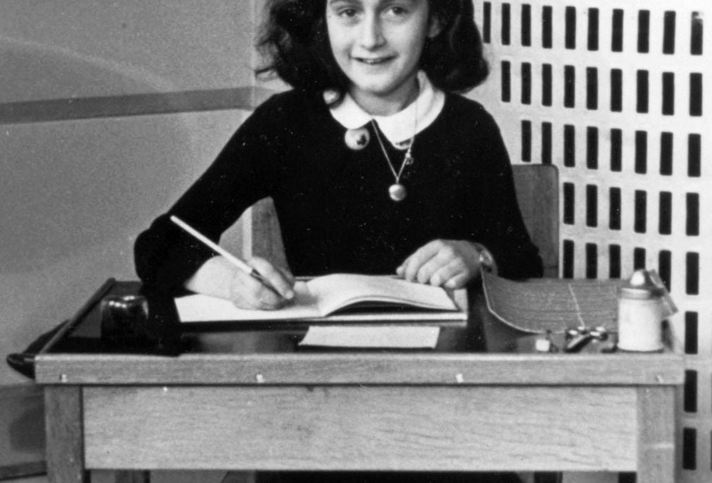 A young Frank sits at a school desk, notebook open and pencil poised to write, smiling and wearing a school uniform