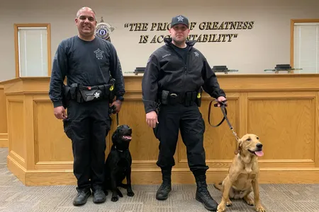 Officers Paul Douglas (left) and Theodore Santos (right) stand with their newest Covid-19 K9 unit: a female black lab named Huntah (left) and a male golden lab-retriever mix, Duke (right).

