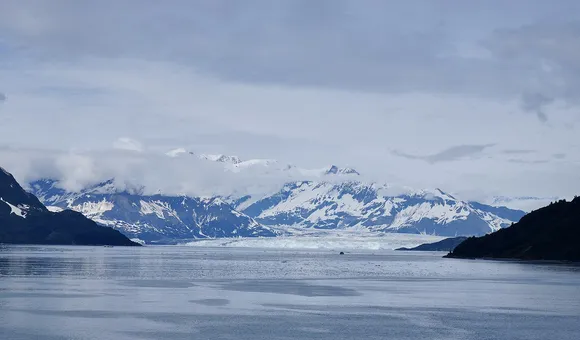 A landscape shot featuring a cloudy gray sky, snow-covered mountains in the distance above a turquoise expanse of glacial ice on the horizon and a dark blue expanse of water in the foreground.