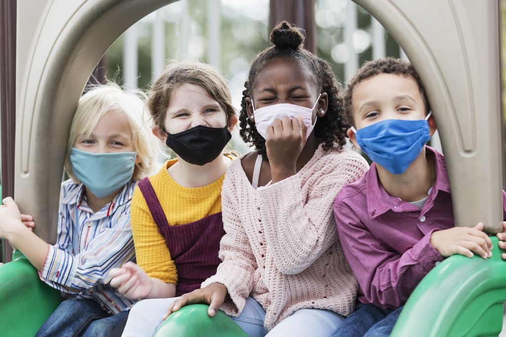 A diverse group of masked children sit at the top of a slide at a park