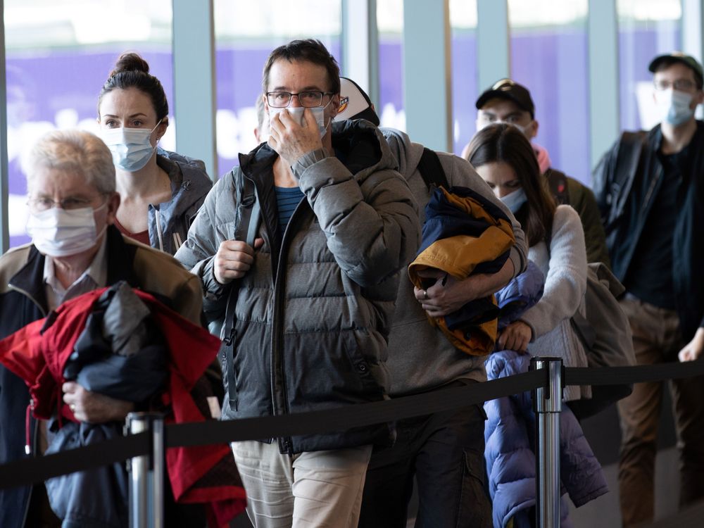 A crowd of people walk through an airport wearing masks. The man in the center of the image is touching his mask with one hand