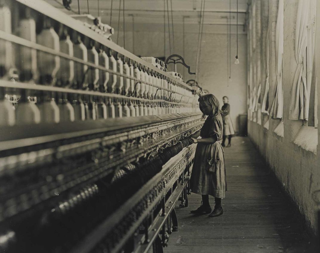 Black and white photo of young girl in a dress, working in a textile mill