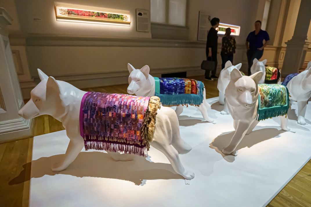 models of dogs wear colorful beaded blankets on their backs in the exhibition