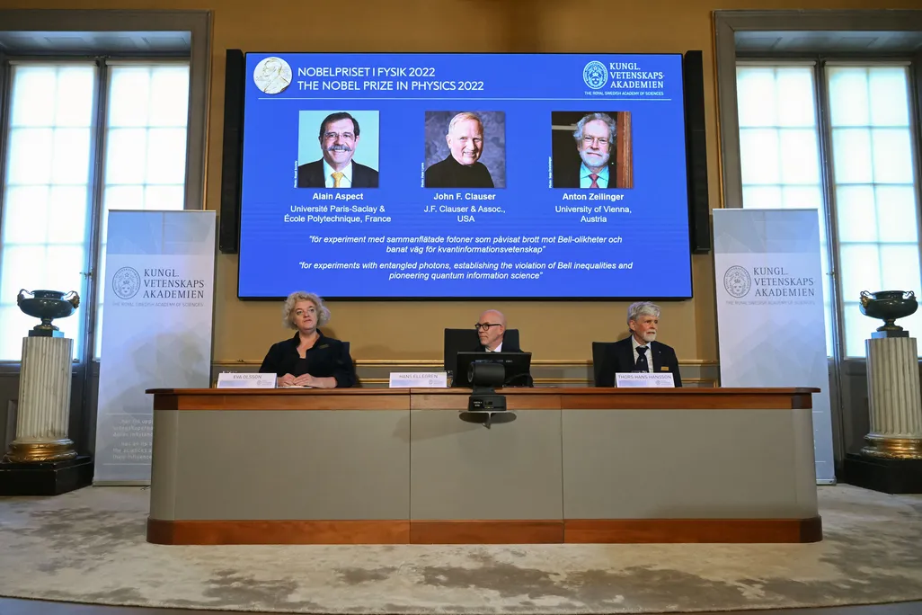 Members of the Nobel Committee for Physics seated at a table, with a screen displaying pictures of the winners of the prize behind them.