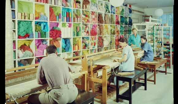 A smiling Dorothy Liebes stands in her studio in her blue apron, supervising two young women sitting at looms with spools of colorful thread and skeins of yarn close at hand.