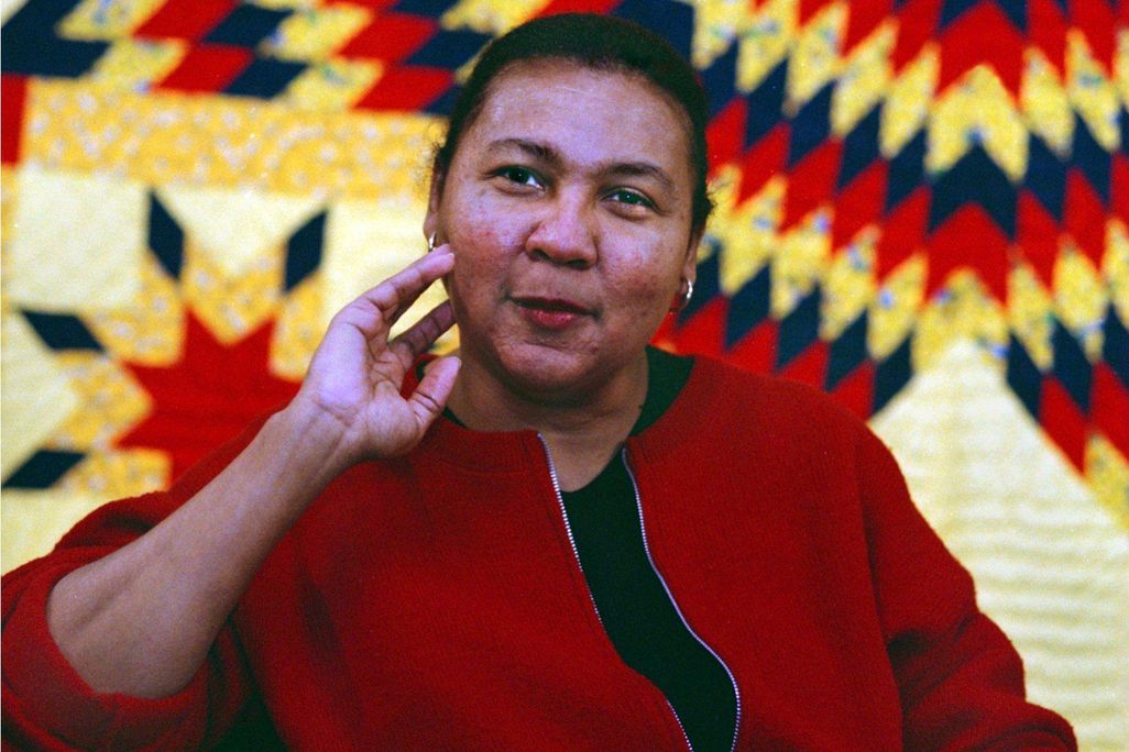 hooks, a Black woman in a red blazer, sits in front of a red, yellow and black quilt with her hand resting lightly on her chin and a small smile