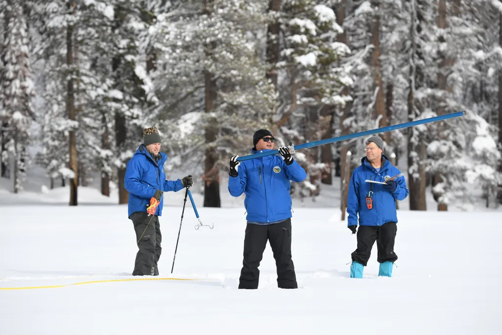 Three men in blue coats standing in the snow with equipment
